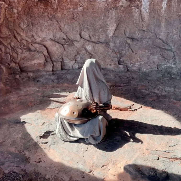 Una figura cubierta por una tela blanca toca un handpan sentada en la arena, dentro de una cueva de roca iluminada por el sol